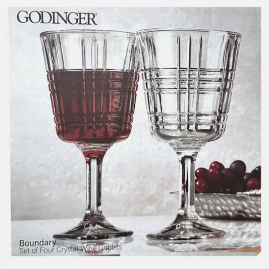 Two crystal wine glasses with red wine and grapes on a textured surface, featuring the brand 'Godinger'.
